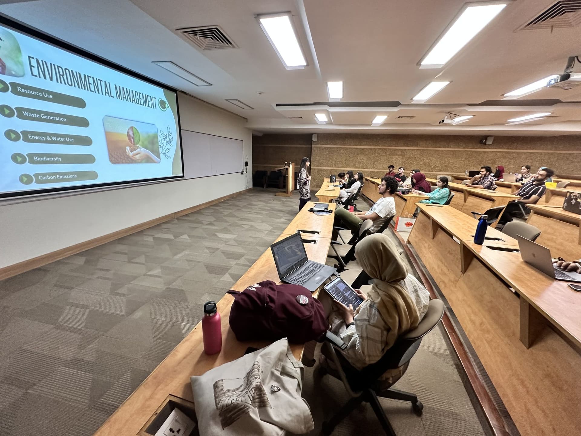 A lecture hall of students facing a screen titled Environmental Management, with Lalain at the front