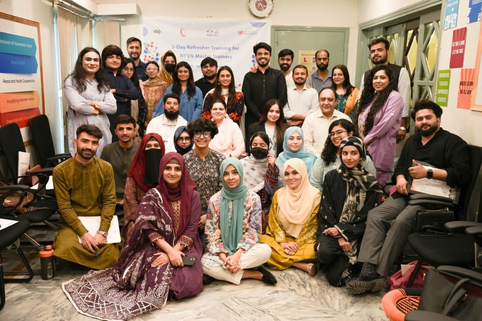A large group of trainers posing in a training room under a banner for the 3-day refresher training