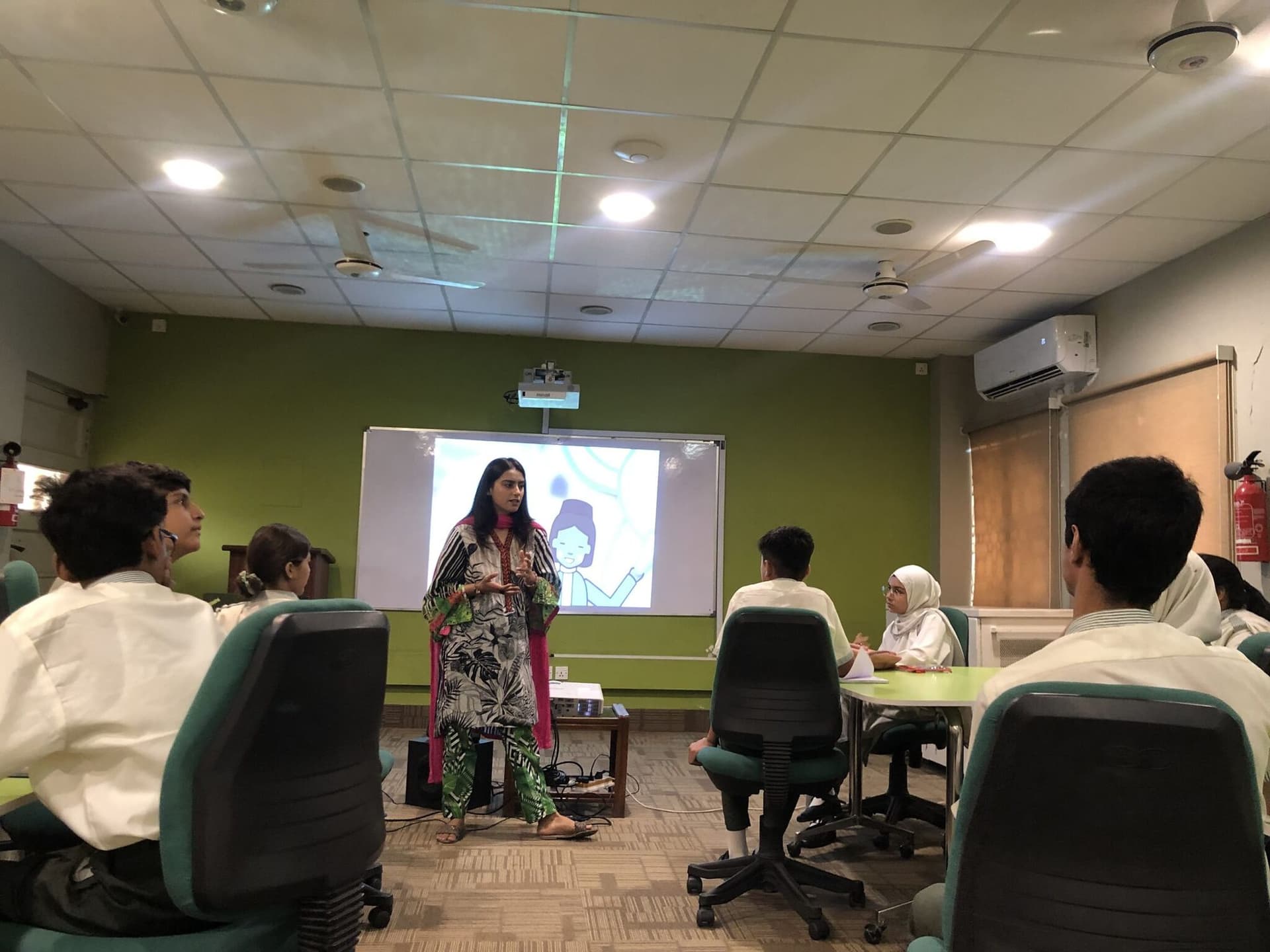 A facilitator at the front of a classroom of students seated at round tables, a slide on the screen behind