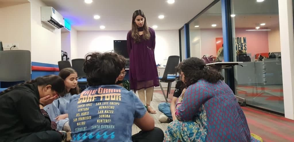 Teenagers sitting on the floor in a circle while Lalain stands and listens